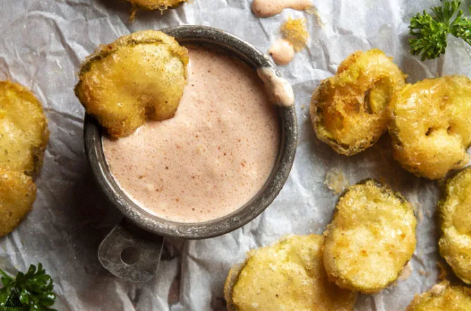 A piece of low carb fried pickles in a bowl of dip with the other low carb fried pickles on the side